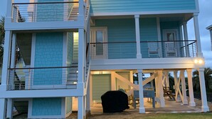 A two-story beachfront house with a balcony, a covered porch, and a small outdoor seating area. - Across The Street From The Beach, Private Beach & Pool Access, Elevator (Holden Beach)