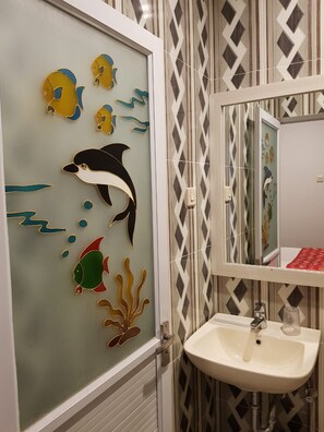 Un baño con puerta de cristal que presenta un mural de delfines, un lavamanos blanco y un espejo con marco de diseño de diamantes.