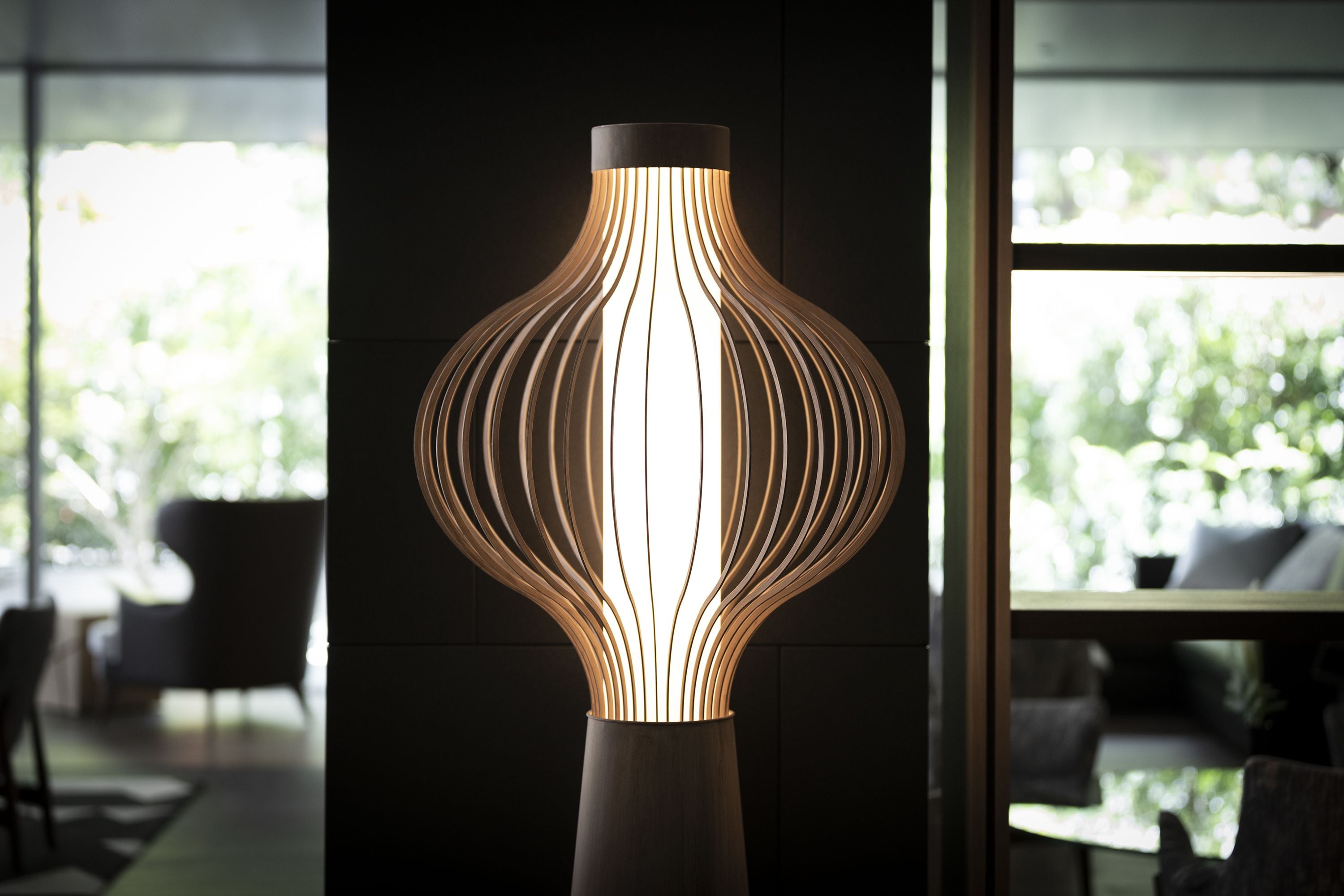 A modern, illuminated floor lamp with a unique, ribbed design in a contemporary room with large windows and a view of greenery outside.