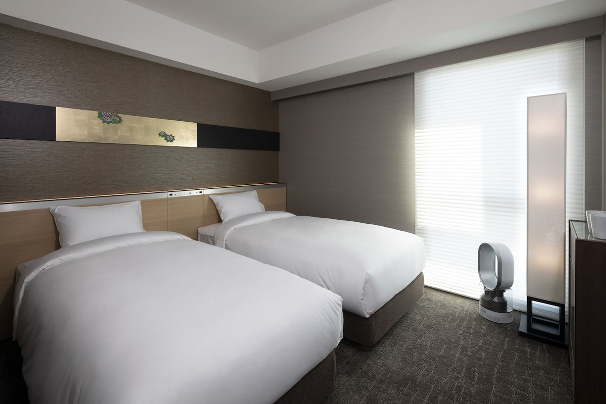 A hotel room with two beds, a large window with blinds, a wall-mounted artwork, and a bedside table with a lamp.
