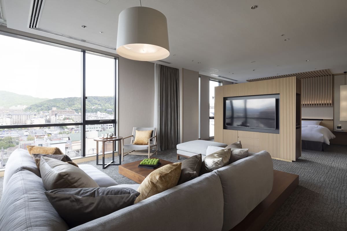 A modern hotel room with a large sofa, a TV, a dining table, and a city view.
