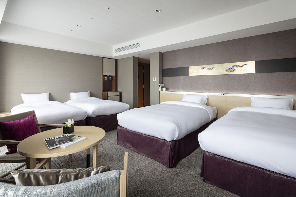 A hotel room with two beds, a small round table with a magazine, a chair, and a decorative wall piece.