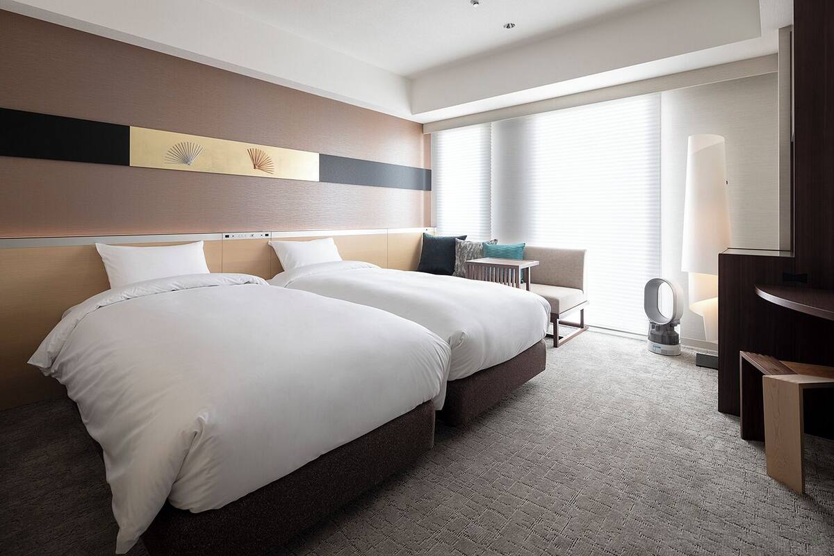A hotel room with two beds, a sofa, a bedside table, a lamp, and a large window with blinds.