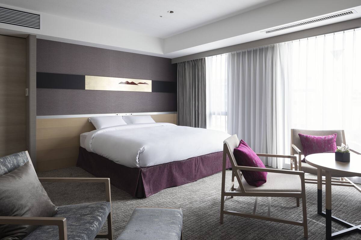 A modern hotel room with a large bed, a desk, two chairs, and a small table.