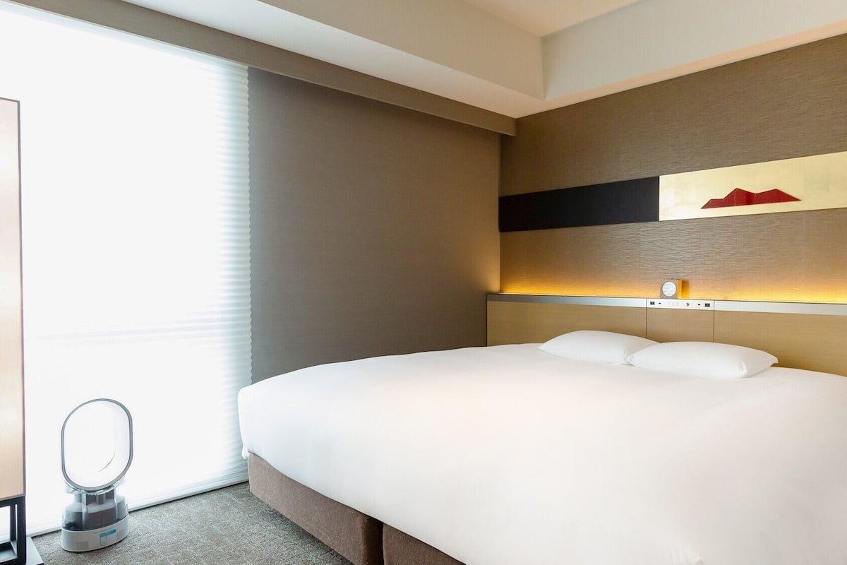 A hotel room with a large bed, a bedside table, a mirror, and a wall-mounted clock.