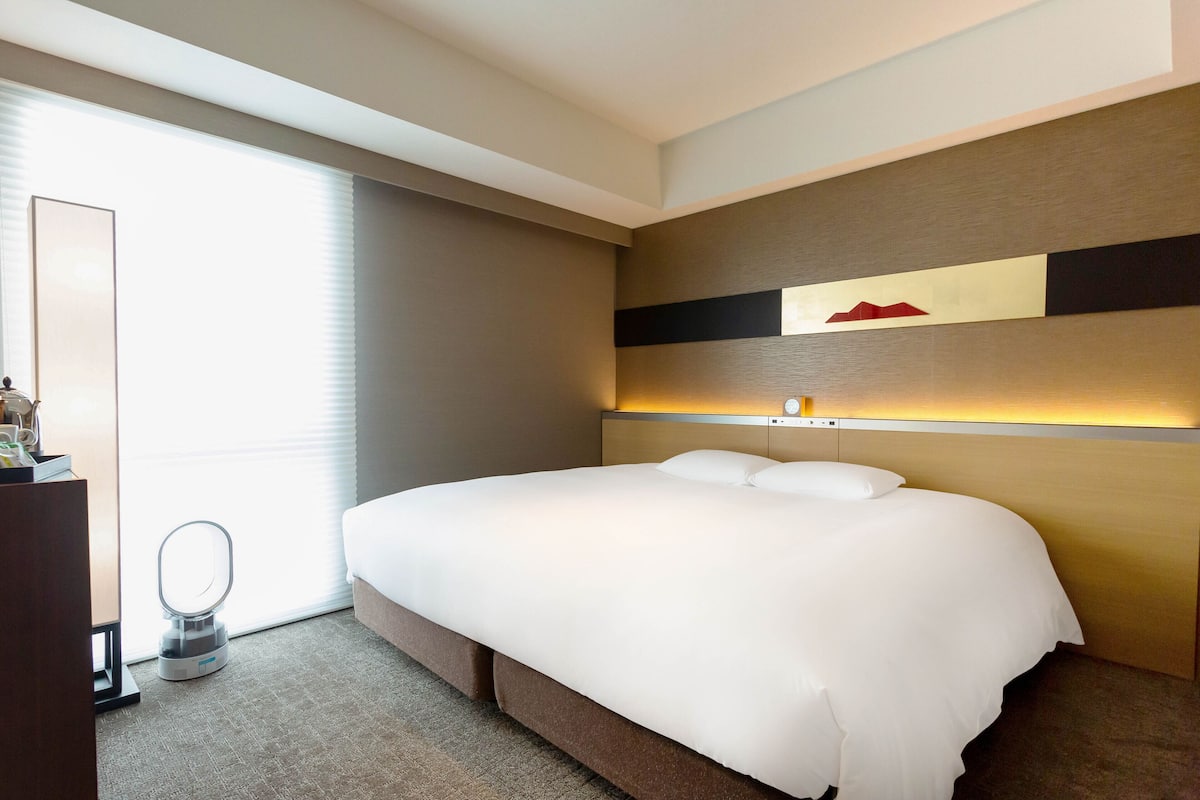 A hotel room with a large bed, a bedside table, a mirror, and a window with blinds.