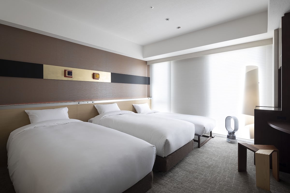A hotel room with two beds, a desk, a chair, and a large window with blinds.