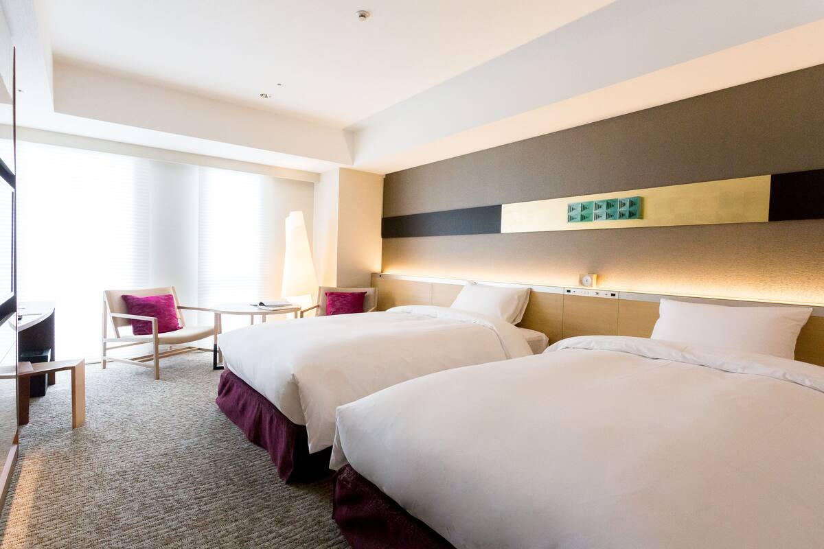 A hotel room with two beds, a desk, a chair, and a large window.