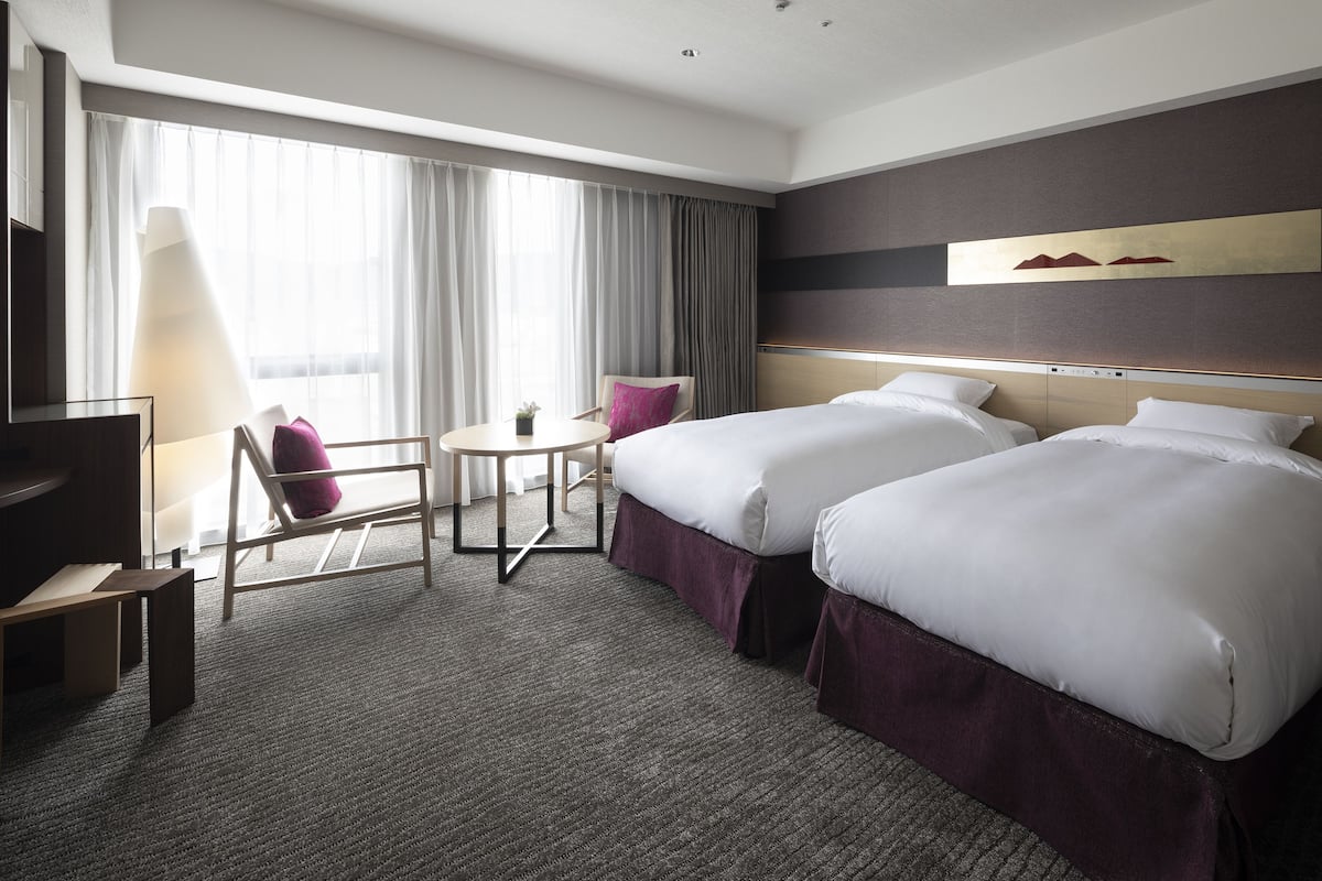 A hotel room with two beds, a desk, a chair, and a large window with curtains.