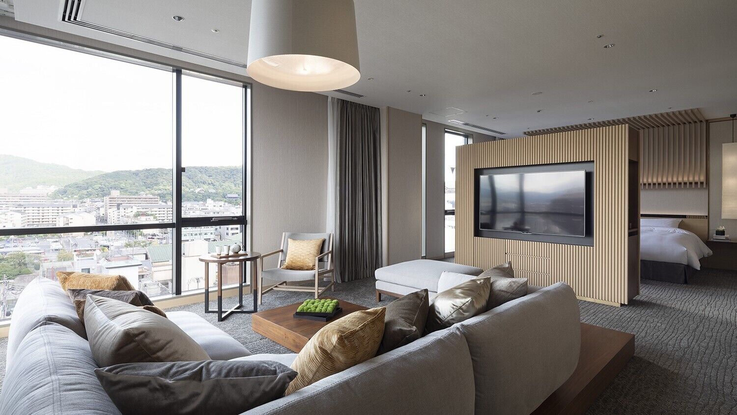 A modern hotel room with a large TV, a sofa, a chair, and a view of the city.