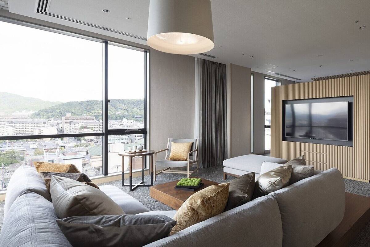 A modern hotel room with a large TV, a sofa, a chair, and a view of the city.