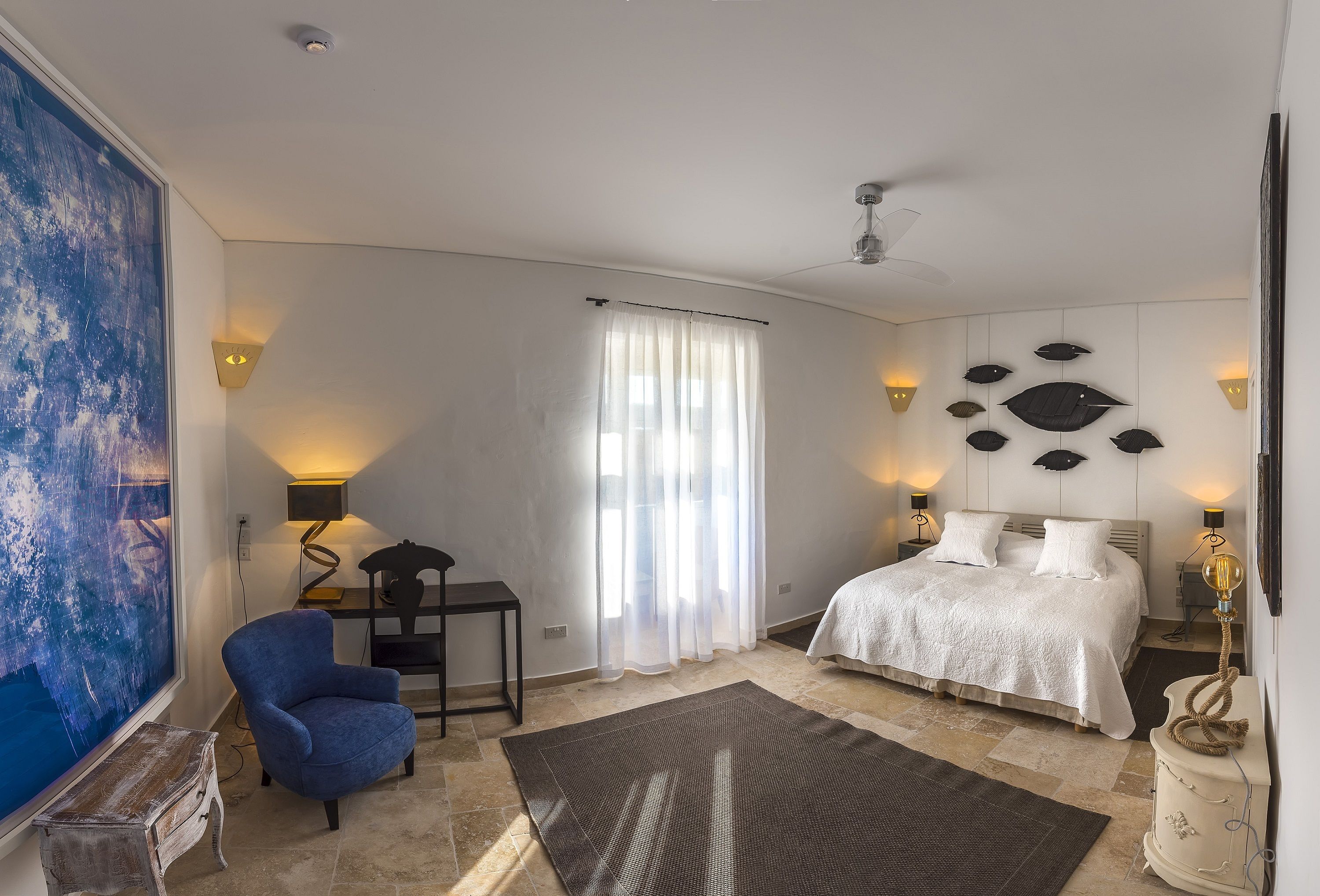 A bedroom with a bed, a desk, a chair, a large painting, and a ceiling fan.