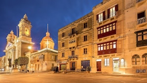 A historic building with a clock tower and a dome, surrounded by other buildings with balconies and storefronts. - The Snop House (Senglea)
