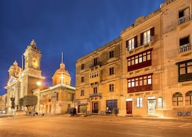 Um edifício histórico com torre do relógio e cúpula, rodeado por outros edifícios com varandas e lojas.