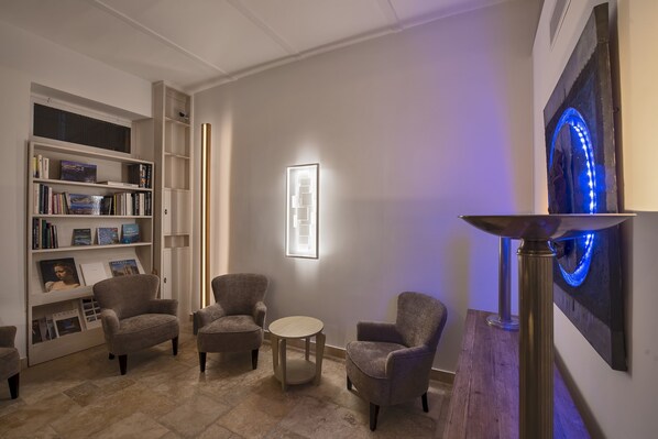 A modern lounge area with a bookshelf, armchairs, a small round table, and a wall-mounted light fixture. - The Snop House (Senglea)