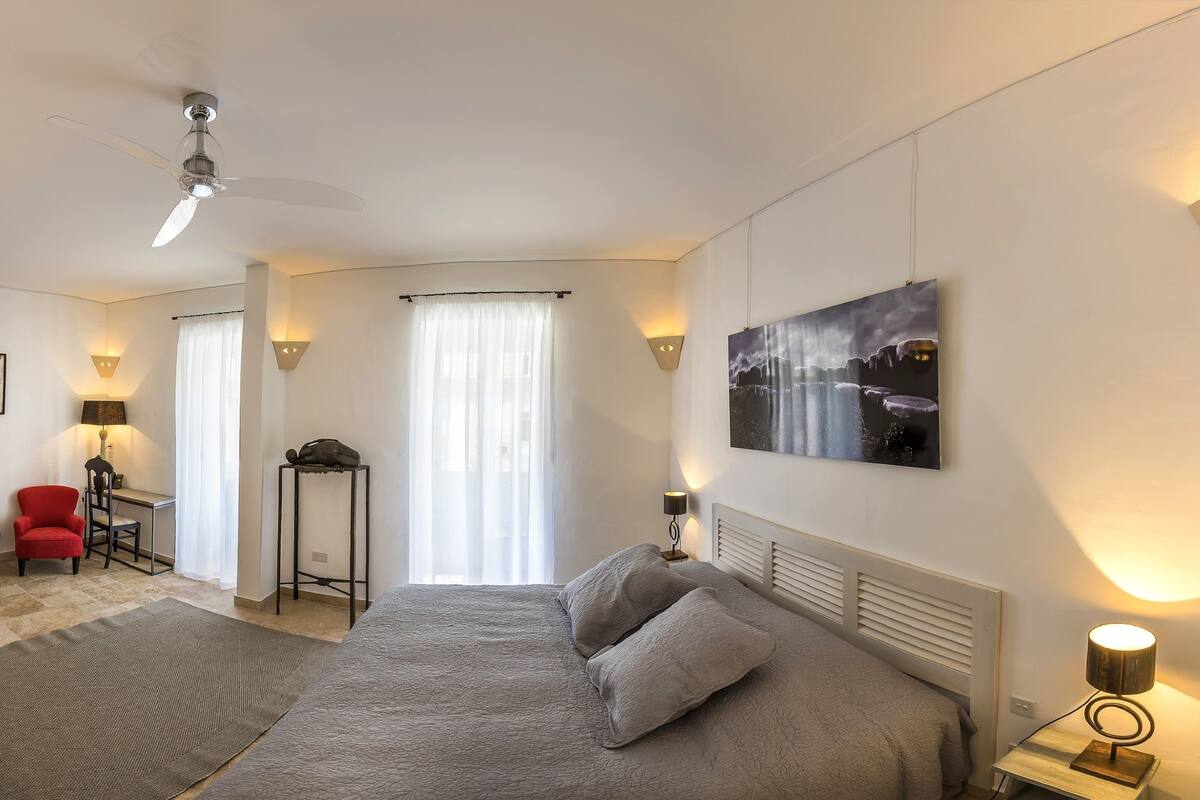 A modern hotel room with a bed, a desk, a chair, a standing lamp, and a ceiling fan.