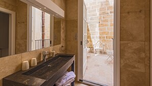 A bathroom with a dark wooden vanity, a large mirror, and a view of a stone patio with outdoor furniture. - The Snop House (Senglea)