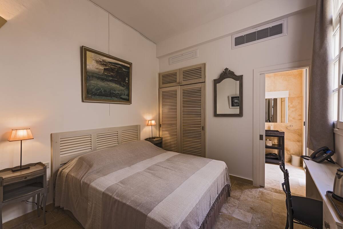 A bedroom with a bed, bedside lamps, a painting, a wardrobe, a mirror, and a desk with a telephone.