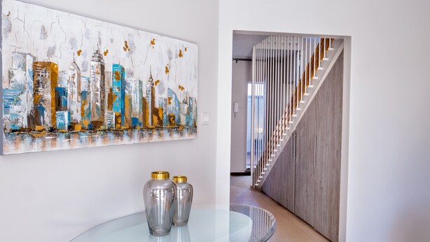 A modern interior with a glass table, a cityscape painting, and a wooden staircase.