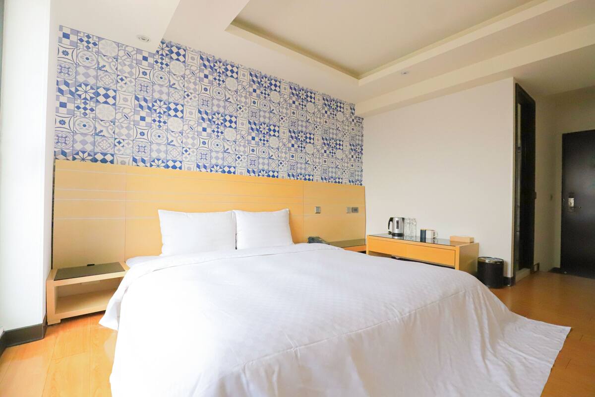Habitación de hotel con una cama grande, una mesita de noche con lámpara, un estante pequeño y una pared con azulejos blancos y azules.