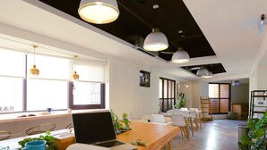 A modern office space with wooden desks, laptops, and books. There are large windows allowing natural light, and the room has a high ceiling with pendant lighting.