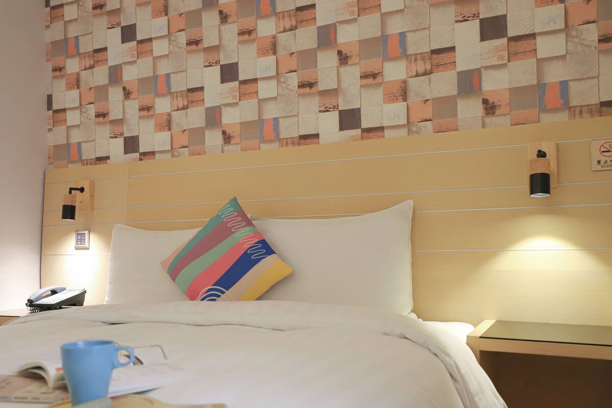 Una habitación de hotel con un estampado de cuadros en la pared, una cama con ropa blanca, una almohada de colores, una mesita de noche con una lámpara y un teléfono en la mesita.