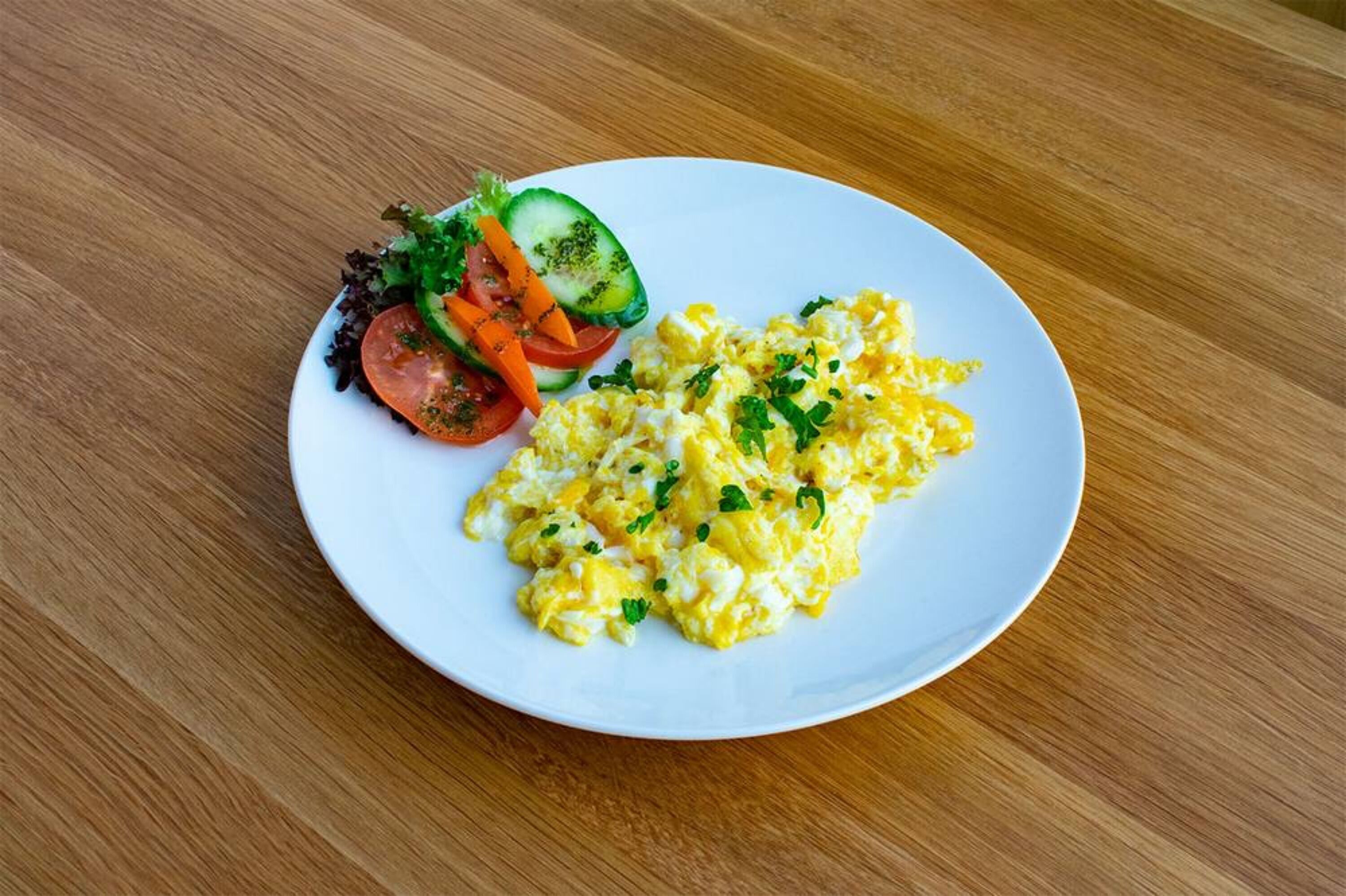 A plate of scrambled eggs with a side salad of lettuce, cucumber, tomato, and carrot.