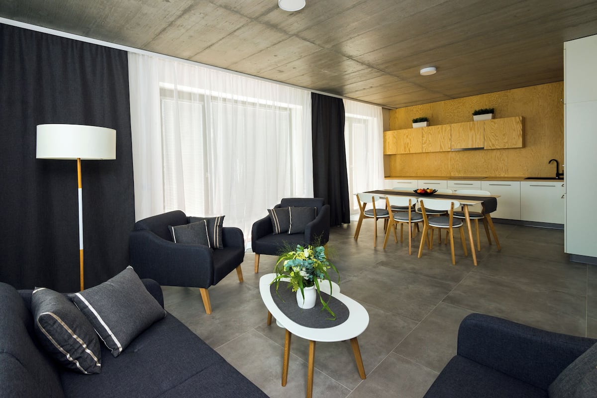 A modern living room with a sofa, armchair, and coffee table, a dining area with chairs and a table, and a kitchen with wooden cabinets.