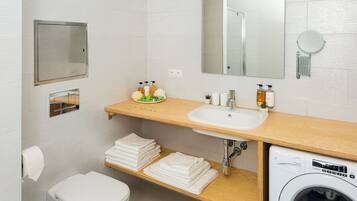 A modern bathroom with a white sink, mirror, and toilet. There is a wooden shelf with towels and toiletries.
