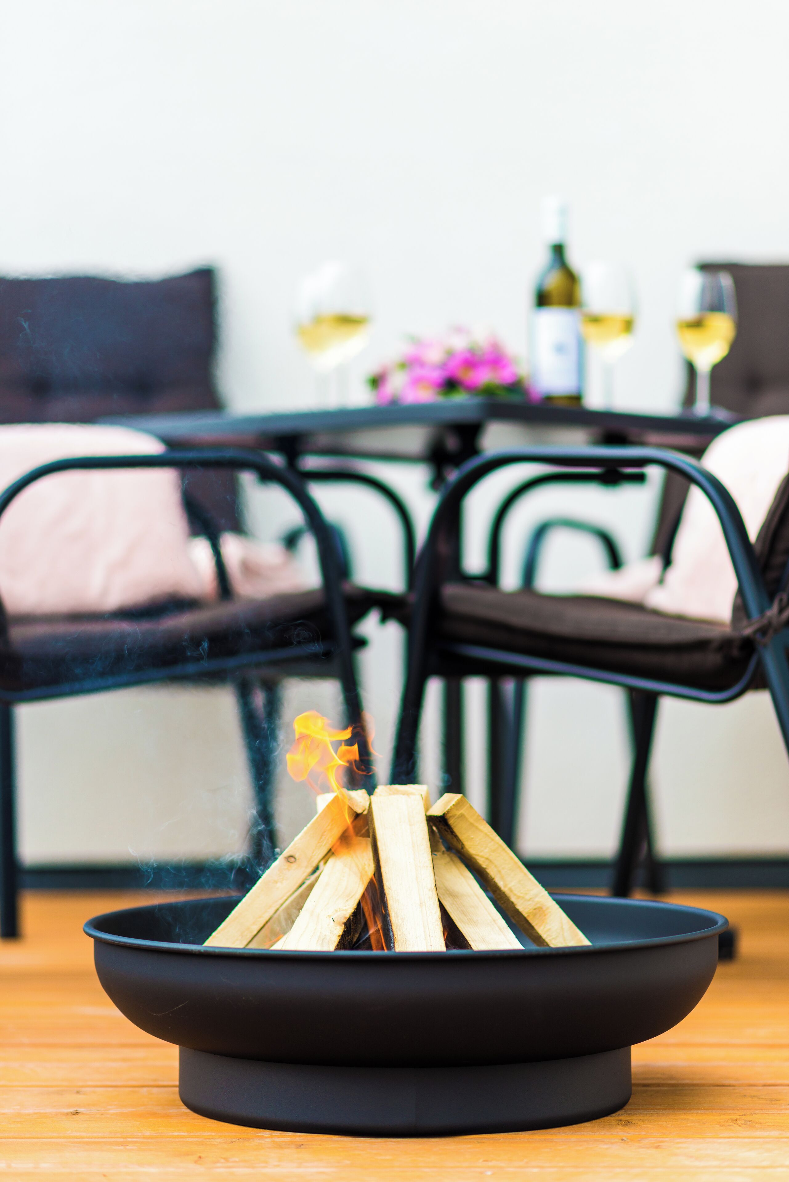 A black fire pit with burning wood, surrounded by a dining table with chairs, and a sofa in the background.