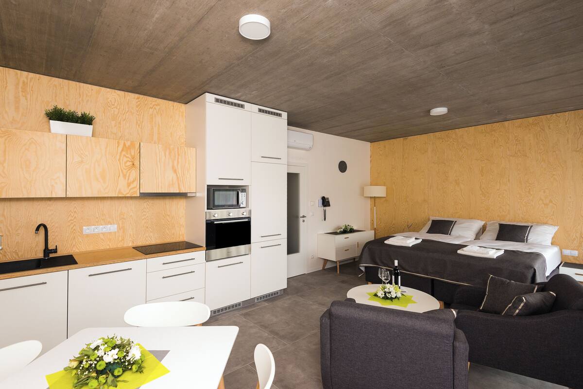 A modern hotel room with a kitchenette, a bed with black bedding, a grey sofa, and a small table with a vase of flowers.