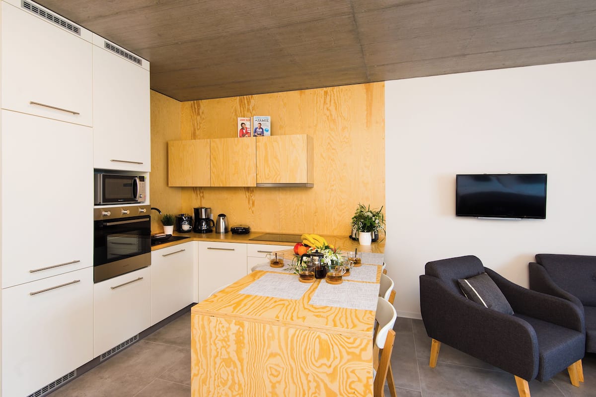 A modern kitchen with white cabinets, a microwave, and a stove. There is a wooden dining table with a placemat and a banana on it. A flat-screen TV is mounted on the wall.