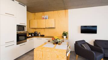 Une cuisine moderne avec des armoires blanches, un four à micro-ondes et une cuisinière. On y trouve une table à manger en bois, avec un set de table et une banane posée dessus. Un téléviseur à écran plat est fixé au mur.