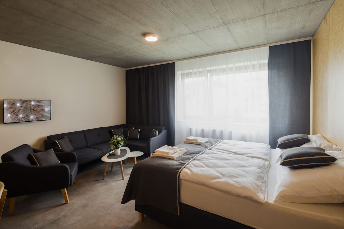 A modern hotel room with a bed, two sofas, a small table, and a large window with curtains.