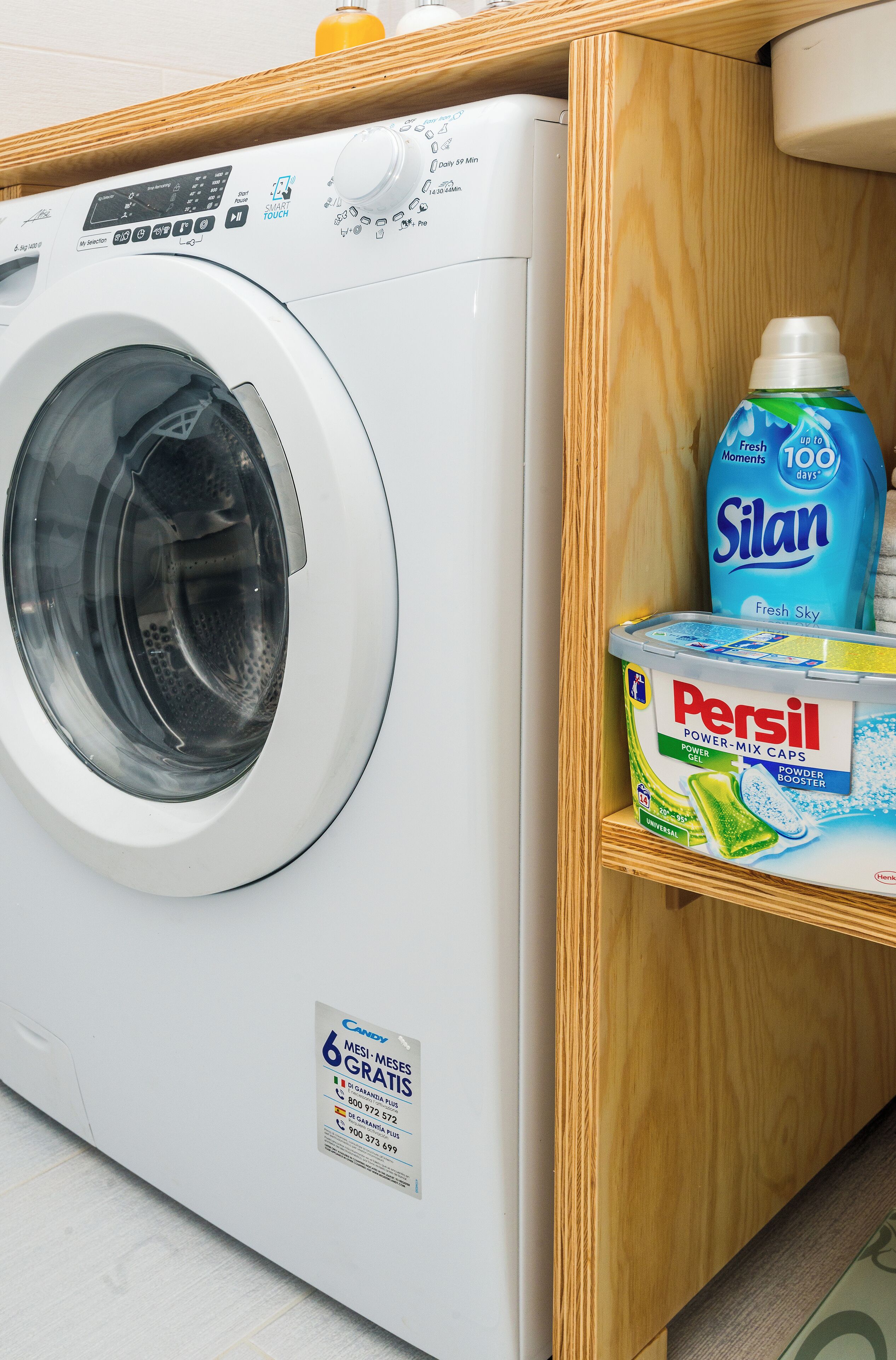 Una lavatrice a carica frontale con pannello di controllo, un contenitore di detersivo Persil e una bottiglia di detersivo Silan.