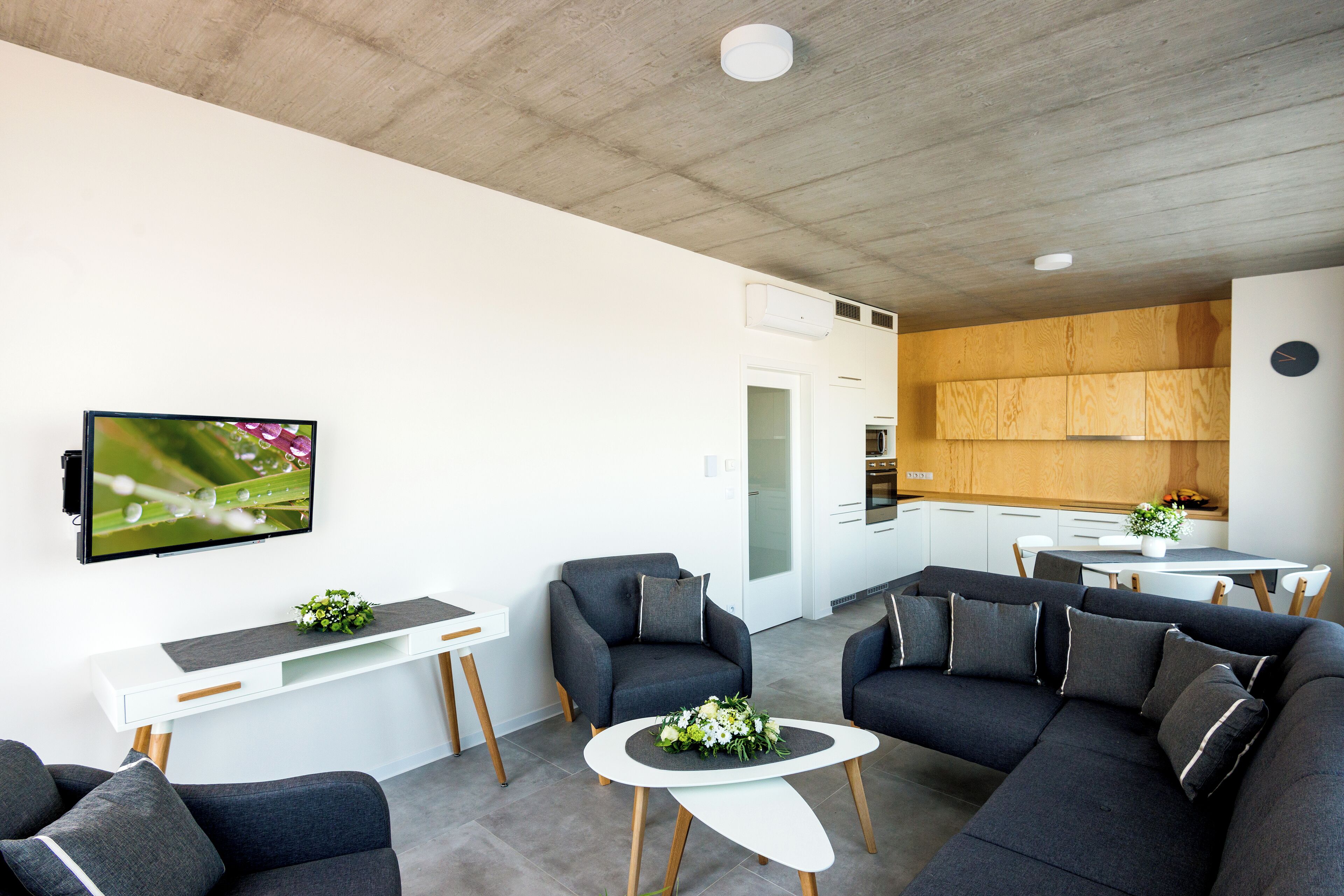 Un salon moderne avec un canapé, des fauteuils, une table basse et un téléviseur fixé au mur. On aperçoit un coin cuisine à l’arrière-plan.
