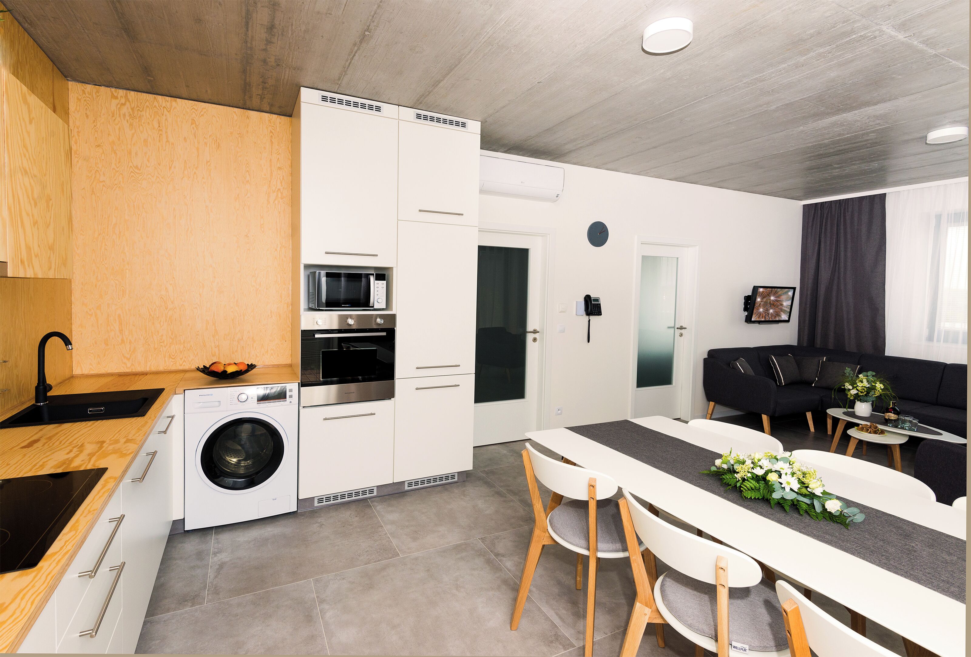 Cuina moderna amb rentadora, microones i forn. Hi ha una zona de menjador amb taula i cadires, i un sofà negre al saló.