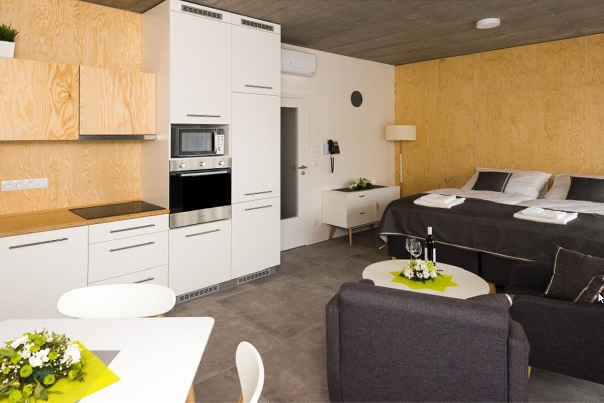 A modern hotel room with a kitchenette, a bed with a black and white bedspread, a grey sofa, and a small dining table with a bouquet of flowers.