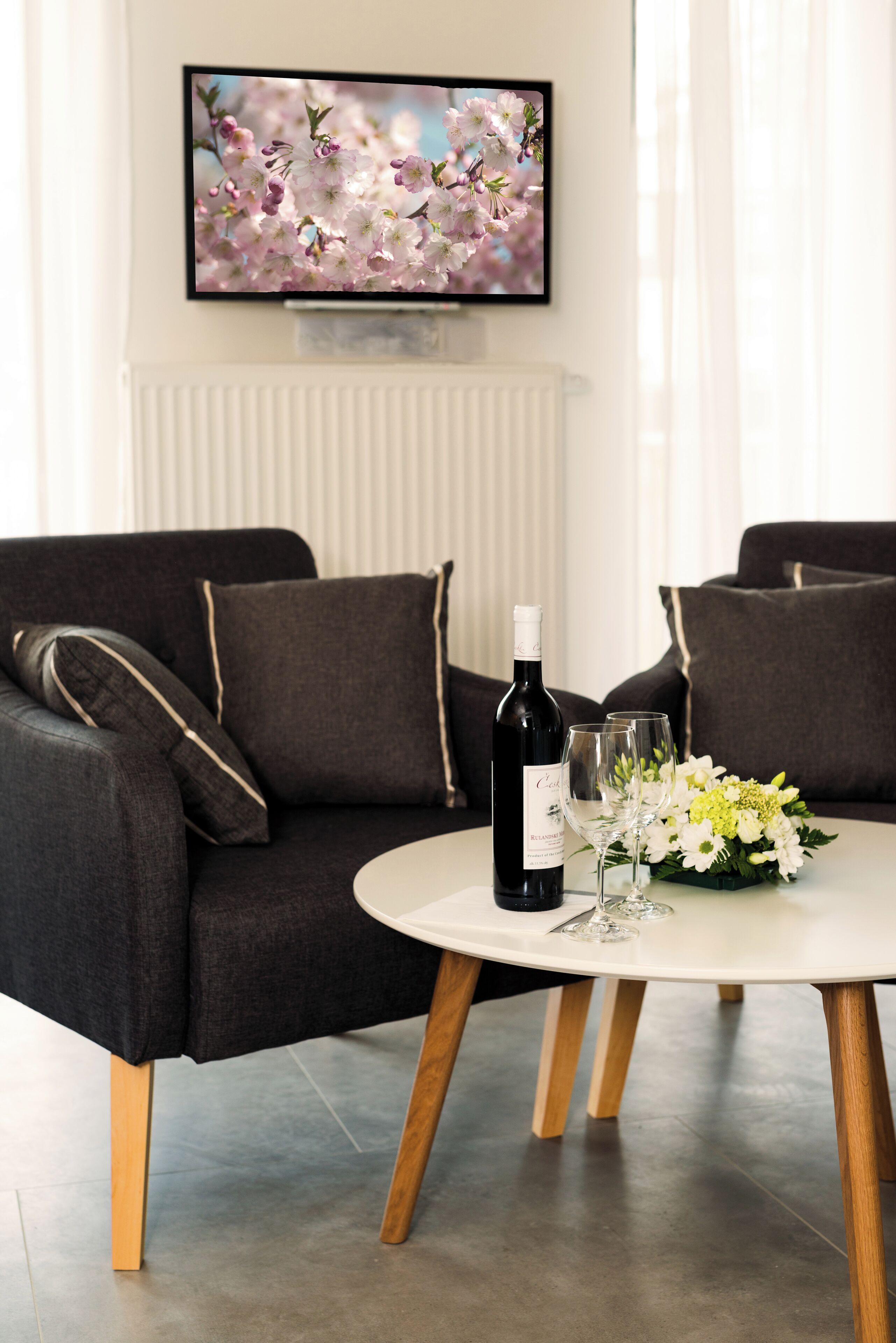 Ein modernes Wohnzimmer mit einem dunkelgrauen Sofa, einem runden weißen Tisch mit einer Weinflasche und Gläsern sowie einer Blumenkomposition.