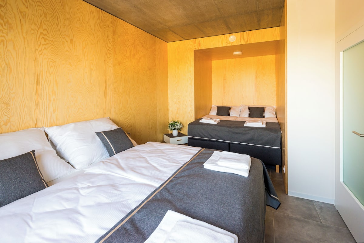 A modern hotel room with two beds, wooden paneling on the walls, and a small bedside table with a plant.
