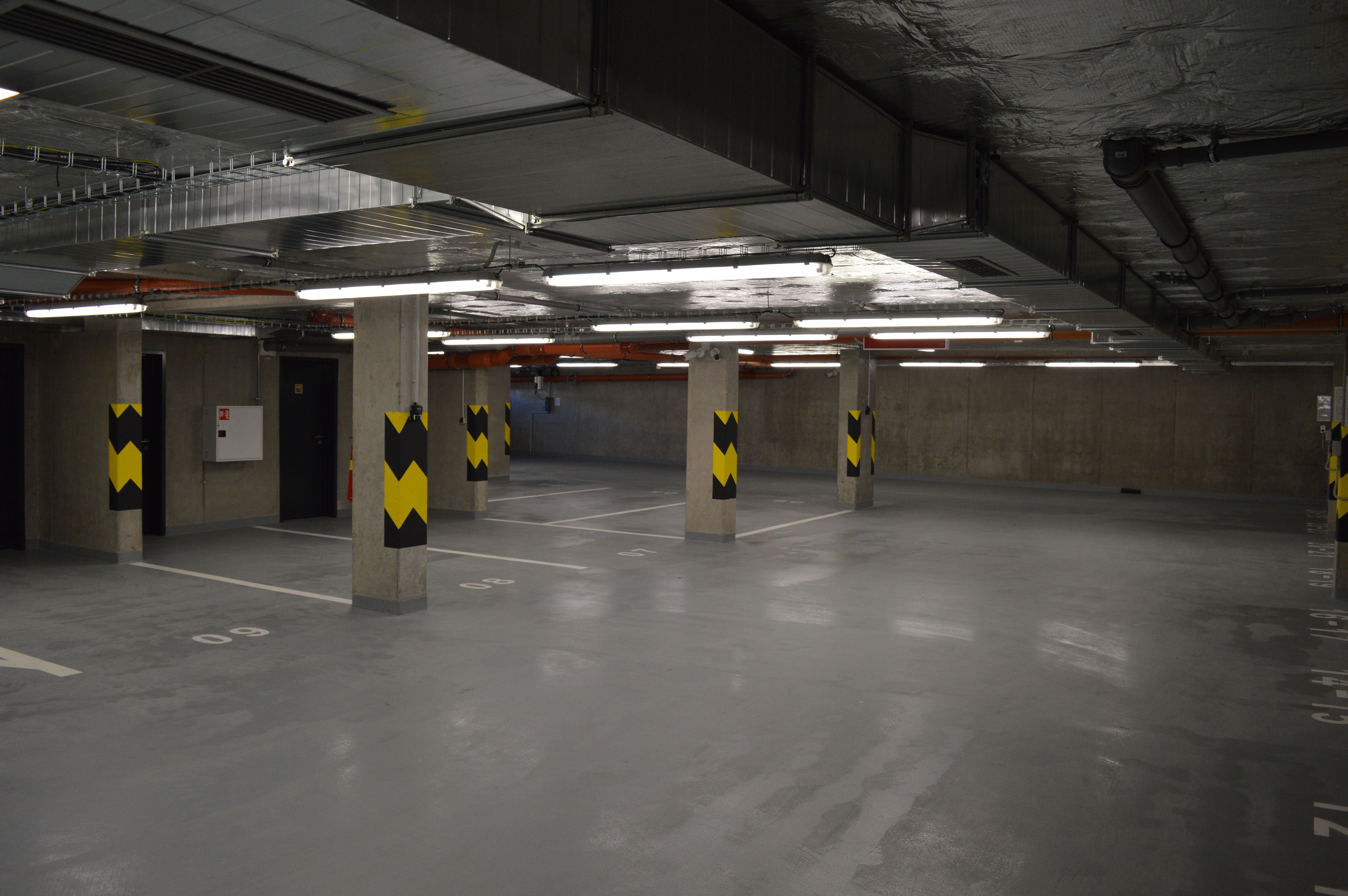 An indoor parking garage with concrete pillars, fluorescent lighting, and marked parking spaces.