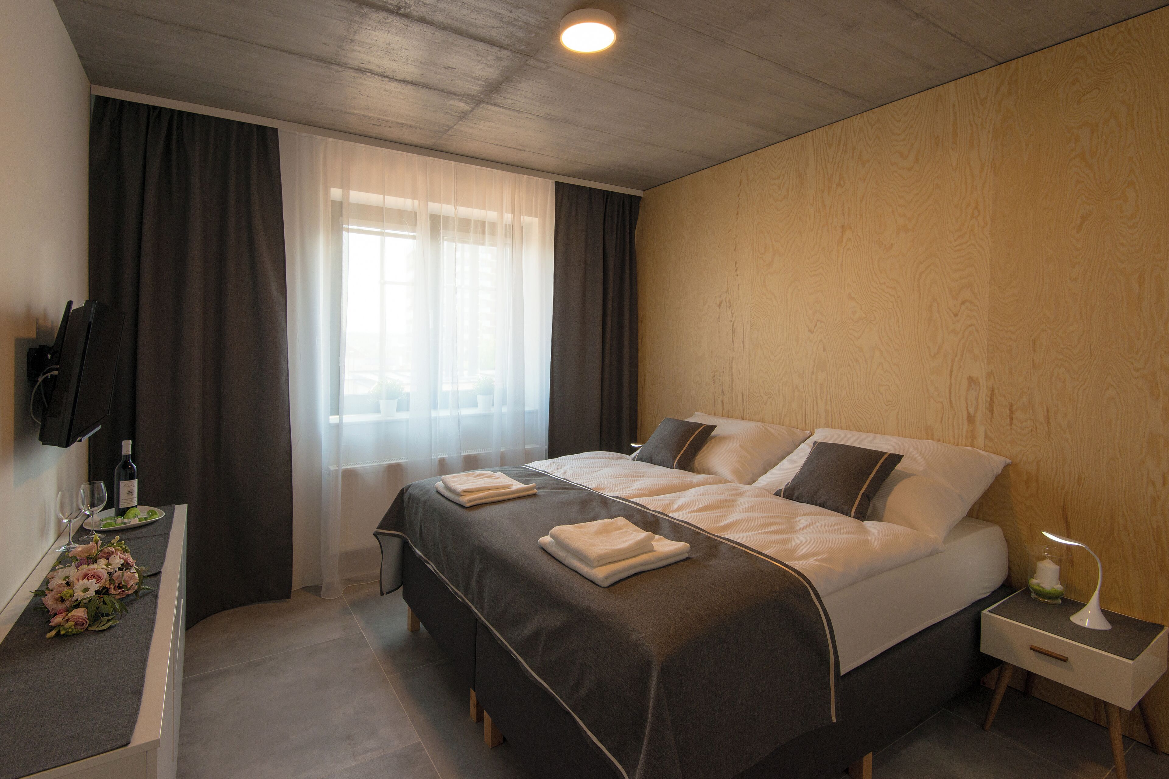 A modern hotel room with a large bed, a bedside table with a lamp, a TV mounted on the wall, and a window with curtains.