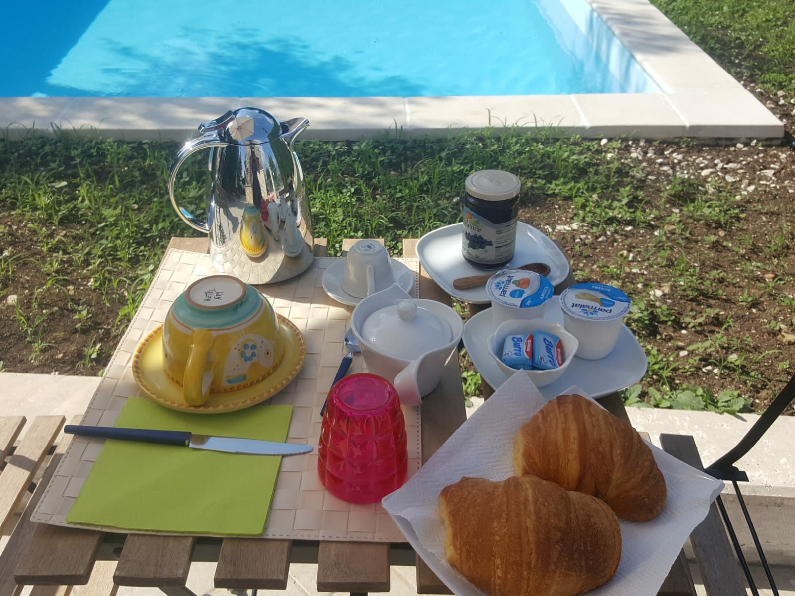 Preparació de l'esmorzar al costat d'una piscina, amb una cafetera, tasses, envasos de iogurt, croissants i un ganivet damunt una taula de fusta.