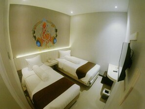 Une chambre d’hôtel avec deux lits, une petite table de chevet avec une lampe, un téléviseur fixé au mur et une petite poubelle.