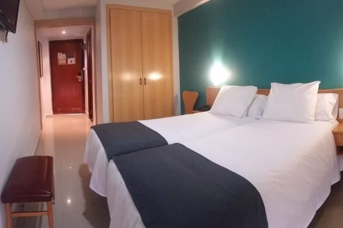 A hotel room with a large bed, a wooden wardrobe, a bench, and a nightstand with a lamp.
