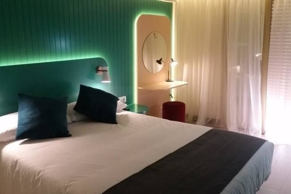 A hotel room with a green headboard, a bed with white and dark blue bedding, a bedside table with a lamp, a mirror, and a window with sheer curtains.