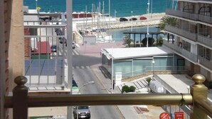 Vista costera con un puerto deportivo, un edificio de varios pisos con balcones y un mar azul y despejado.