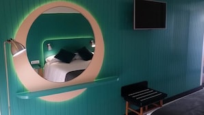 Una habitación con un espejo circular en la pared, una cama con almohadas, una lámpara de noche y un banco negro.