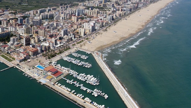 Vista aérea de una ciudad costera con un puerto deportivo, una extensa playa de arena y edificios urbanos densos.