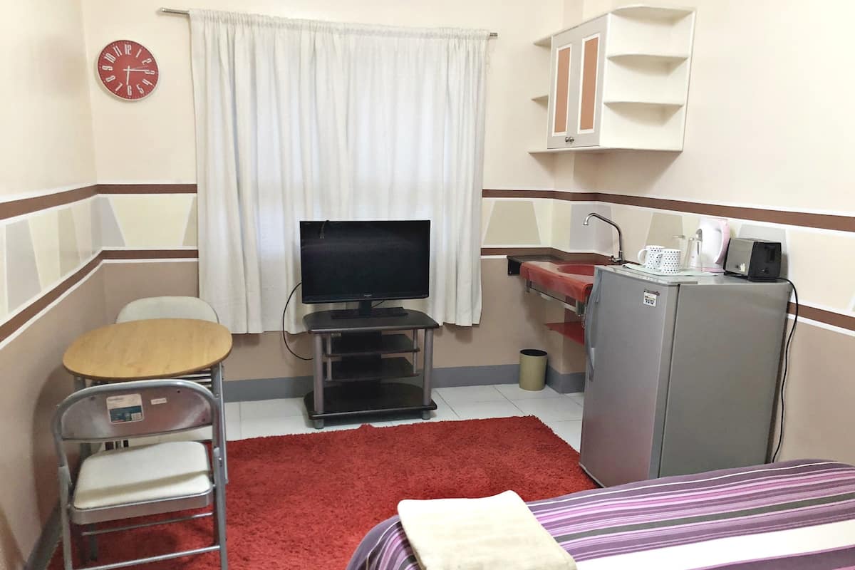 A hotel room with a bed, a desk with a chair, a television, a small kitchenette, and a red carpet.